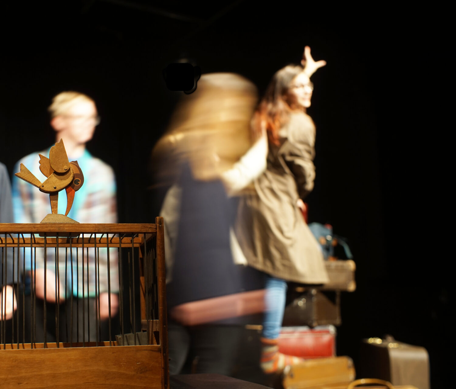 Ein Vogel sitzt auf einem Käfig. Im HIntergrund spielen Kinder Theater.